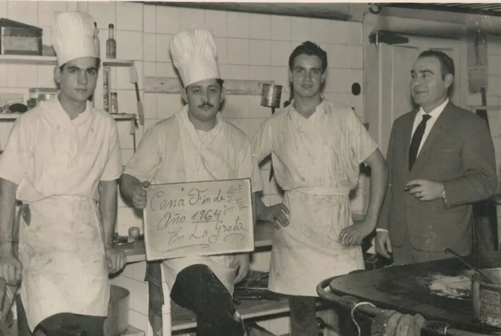 Fotografía de archivo familiar de 1964: cocineros y un hombre con traje en una cocina, con un cartel que dice Cena Fin de Año 1964 en La Gruta
